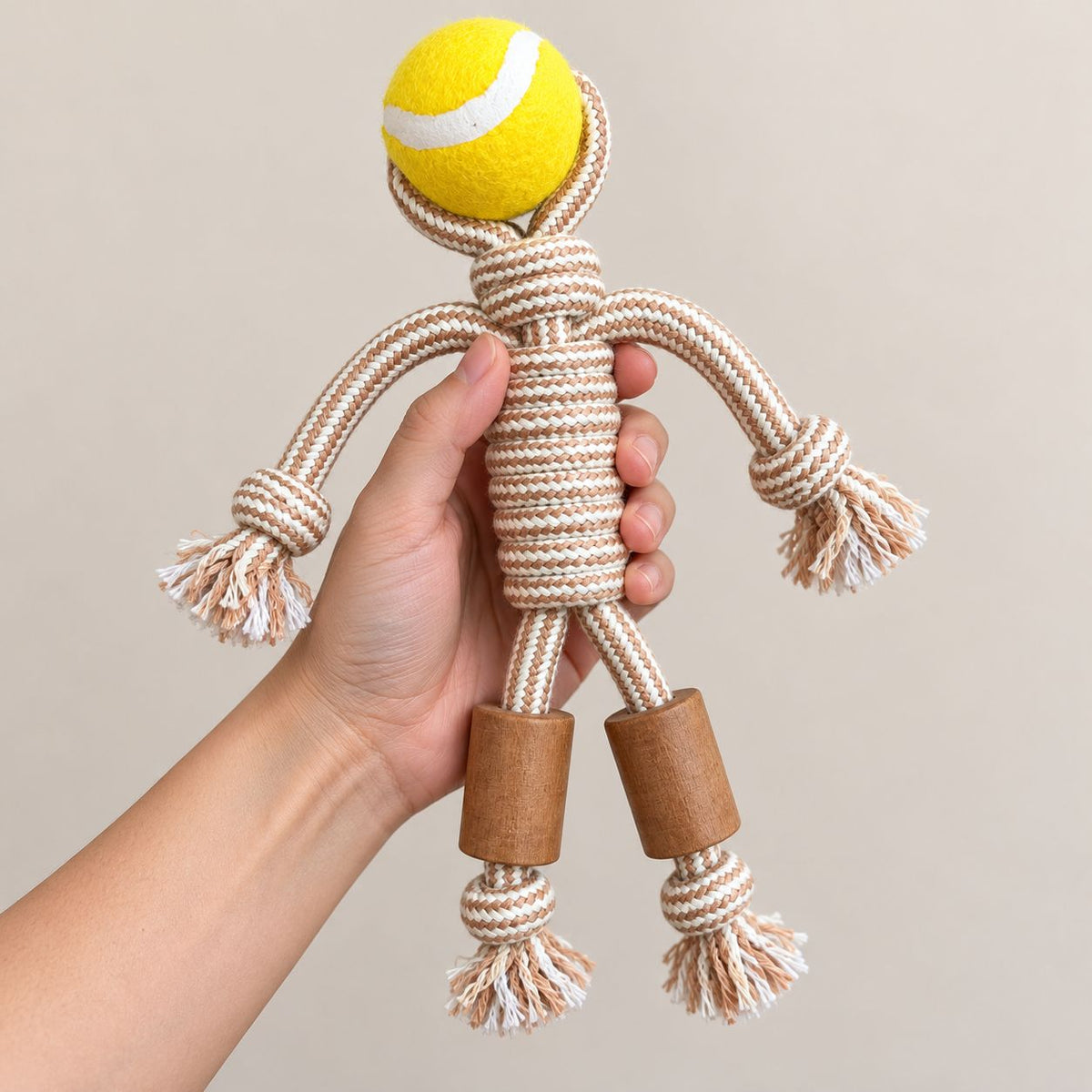 Hand holding a rope dog toy with a yellow tennis ball head and wooden legs.